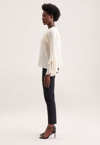 Blusa color crema con maniche a volant, abbinata a jeans neri attillati e tacchi a spillo neri. Tessuto liscio con un design fluido.