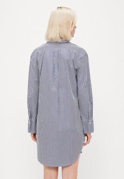 Pessoa loira de costas, vestindo um vestido camisola azul e branco com riscas verticais e punhos abotoados.