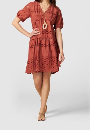 Femme portant une robe à œillets couleur rouille, longueur genoux, avec manches bouffantes, ourlet festonné, un long collier pendentif et des sandales.