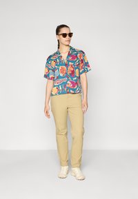 Maglietta a maniche corte di colore turchese con motivi di pesci colorati; indossata con pantaloni beige e sneakers chiare. Modello con occhiali da sole.