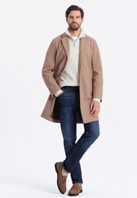 Abrigo de lana beige con solapa de muesca, superpuesto a un suéter de punto gris claro. Jeans azul oscuro y mocasines marrones completan el conjunto.