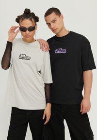 Two individuals are wearing oversized T-shirts; one in light gray with a black graphic logo, the other in black with a purple logo.