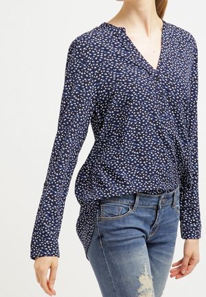 Frau trägt eine marineblaue Bluse mit langen Ärmeln und kleinen weißen und blauen Punkten, kombiniert mit zerrissenen blauen Jeans.