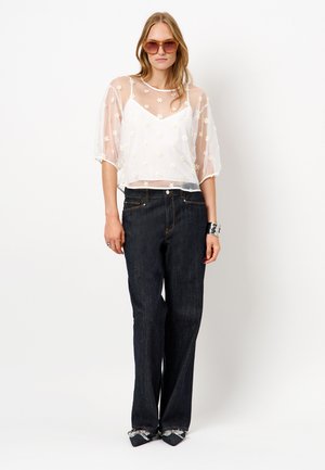 Witte doorschijnende top met bloemachtige borduursels, gecombineerd met donkerblauwe wijde jeans. Model draagt bruine zonnebril en zwarte schoenen met decoratieve accenten.