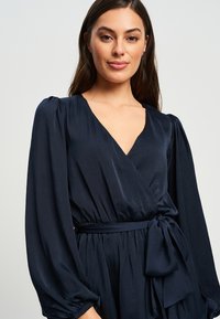 Mornarska obleka z zavihki z dolgimi napihnjenimi rokavi, V-izrezom in detajlom s povezovanjem v pasu. Mehka, lahkotna tkanina z gladko teksturo.