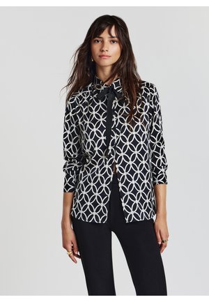 Camicia a righe bianco e nero con un design geometrico, colletto e chiusura frontale con bottoni. Maniche lunghe con polsini abbottonati.