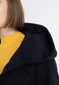 Cappotto in lana blu navy con un ampio cappuccio e colletto asimmetrico, indossato sopra un dolcevita giallo strutturato. Tessuto liscio con linee pulite.