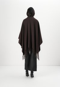 Braune gestrickte Poncho mit Fransen, ausgestattet mit einer Kapuze und einem lockeren Schnitt, getragen über schwarzen Lederhosen und Absatzstiefeln.