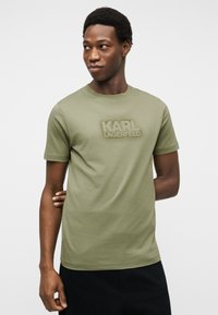 Jeune homme portant un T-shirt Karl Lagerfeld vert olive, debout avec un bras derrière le dos, regardant vers sa gauche.