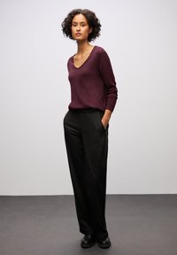 Maglione bordeaux a scollatura a V, maniche lunghe, abbinato a pantaloni neri con tasche laterali. Modello in piedi su uno sfondo neutro.