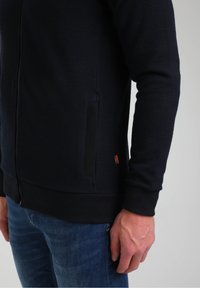 Giacca blu navy da uomo con zip in tessuto testurizzato, presenta un colletto alto, polsini a coste e tasche laterali. Piccolo marchio sul fondo.