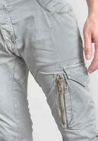 Pantalons cargo gris clair avec une poche zippée bien visible sur la cuisse, des accents cousus et une texture de tissu lisse.
