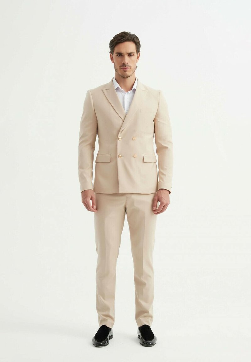 Costume beige à double boutonnage avec des boutons dorés, coupe ajustée, associé à une chemise blanche et des chaussures noires à enfiler. Texture de tissu lisse.