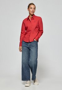 Camicia rossa a maniche lunghe con colletto, indossata con jeans a gamba larga blu e sneakers chiare, con un piccolo emblema sul petto.