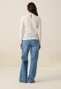 Cremfarbene Spitzenbluse mit langen Ärmeln, Details mit Schlüsselloch-Ausschnitt auf der Rückseite, kombiniert mit hellblauen Schlagjeans. Weiche Textur, durchgängiges Blumendesign.