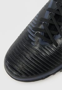 Fekete labdarúgócipő, textúrázott, bordázott felsőrésszel, fekete fűzőkkel és sima orrésszel, amelyet a pályán való tapadásra és irányításra terveztek.