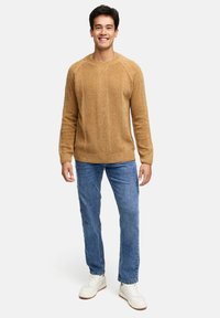 Smilende mand iført en struktureret tan sweater, blå jeans og hvide sneakers, stående mod en ensfarvet lys baggrund.