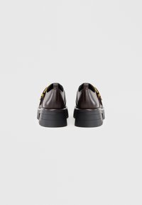 Pruunid nahast kingad koos jämeda musta tallaga ja kollaste aktsentdetailidega. Sile tekstuur, ümar varvas ja madala kaelusega disain.