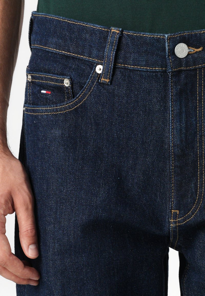 Dunkelblaue Jeans aus Denim mit gelben Nähten, Fünf-Taschen-Design und ein Metallknopfverschluss. Minimale Markenkennzeichnung an der Vorderseite der Tasche.