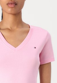 Woman wearing a light pink V-neck T-shirt with a small red, white, and blue rectangular logo on the chest.