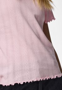 Κοντινό πλάνο ενός ατόμου που φοράει ένα ροζ ανοιχτό πουλόβερ με κοντά μανίκια, με υφασμάτινο μοτίβο και βολάν στο κάτω μέρος, συνδυασμένο με μαύρο παντελόνι.