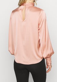Blusa di raso rosa chiaro con colletto alto, apertura a chiave sulla schiena e maniche lunghe caratterizzate da polsini arricciati per texture e dettaglio.