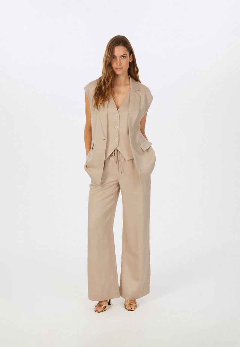 Beige Dreiteiler, bestehend aus einem ärmellosen Weste mit Knöpfen, einer Hose mit Kordelzug und offenen Schuhen mit Absatz, die eine weiche Textur aufweisen.