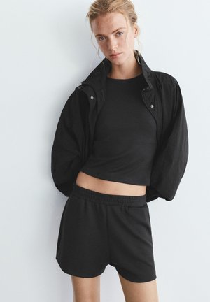 Žena má na sebe čierny krátky top, voľnú čiernu bundu so zvýšeným golierom a čierne elastické šortky na jednoliatom svetlom pozadí.