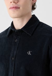 Camicia di velluto a coste nera con colletto, dotata di chiusura frontale con bottoni e logo ricamato sul petto. Tessuto testurizzato con coste visibili.