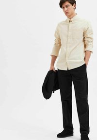 Beige button-up shirt with long sleeves and chest pocket, paired with black trousers and black shoes, holding a black jacket.