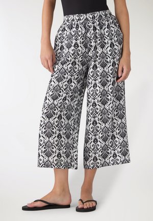 Persona che indossa pantaloni a gamba larga a fantasia bianco e nero e infradito nere, in piedi contro uno sfondo chiaro e uniforme.