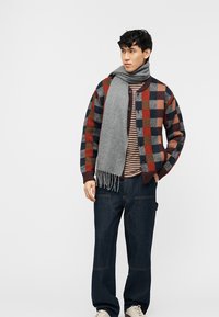 Νεαρός άνδρας που φοράει πολύχρωμο καρό σακάκι, ριγέ πουκάμισο, γκρι κασκόλ, σκούρα τζιν και λευκά αθλητικά παπούτσια, στέκεται με το ένα χέρι στην τσέπη.