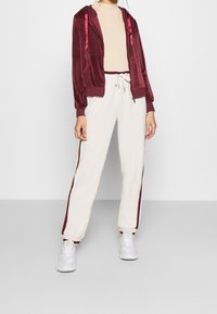 Sudadera de terciopelo burdeos con cremallera y cordones de satén, combinada con pantalones crema ajustados con rayas burdeos a los lados y cintura elástica.