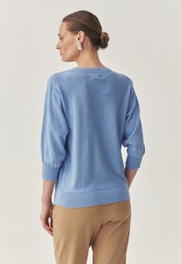 Pulover de culoare albastru deschis, cu mâneci trei sferturi, tiv ondulat și textură moale. Modelul poartă pantaloni bej și cercei argintii.