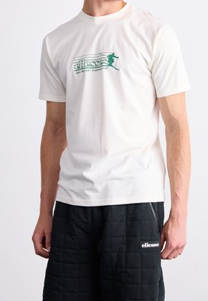 Mężczyzna ubrany w białą koszulkę z zielonym logo narciarskim i napisem, w połączeniu z czarnymi pikowanymi szortami z białą naszywką z logo.