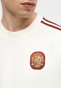 Gros plan d'une personne portant une chemise blanche avec des rayures rouges à l'épaule et un écusson brodé rouge et or détaillé.