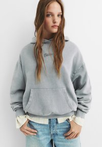 Sweatshirt à capuche gris avec une poche frontale et des poignets côtelés, superposé à une chemise blanche rayée, associé à un jean léger bleu déchiré.