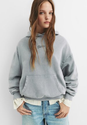Felpa grigia con cappuccio e tasca frontale, con polsini a costine, indossata sopra una camicia bianca a righe, abbinata a jeans chiari strappati.