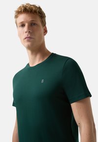 Młody mężczyzna z kręconymi blond włosami, ubrany w ciemnozielony t-shirt z krótkim rękawem z małym logo "B" na piersi, na jednolitym tle.