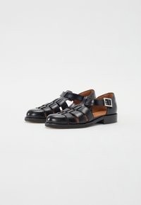 Black leather sandals with a closed-toe design, featuring crisscross straps and a side buckle. Low heel with a textured sole.