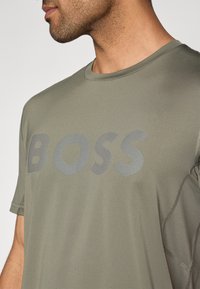 T-shirt atlético verde com um destacado logótipo "BOSS" em riscas horizontais cinzentas. Mangas curtas, decote redondo e textura suave.