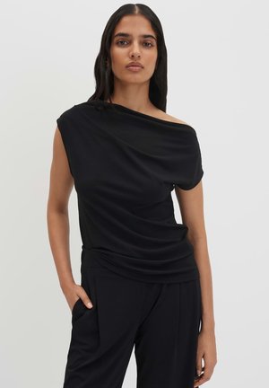 Femme portant un haut noir asymétrique décolleté épaule et un pantalon noir, debout avec une main dans la poche devant un fond clair uni.