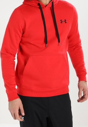 Mann trägt einen roten Under Armour Hoodie mit schwarzen Kordeln und schwarzen Under Armour Hosen, steht mit entspannten Armen.