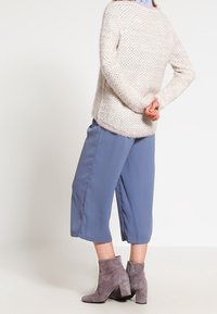 Persona vistiendo un suéter de punto crema, pantalones anchos recortados azules y botas de ante gris hasta el tobillo, de pie con las manos entrelazadas detrás de la espalda.