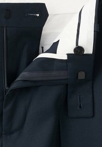 Gros plan d'une ceinture de pantalon bleu marine foncé montrant une fermeture éclair ouverte, un bouton, une doublure intérieure blanche et des détails de boutonnière.
