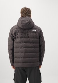 Zwarte pufferjas met capuchon, voorzien van horizontale stiksels, een logo op de linker schouder en een gladde, geïsoleerde textuur.