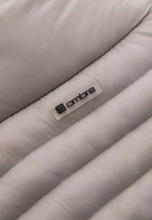 Lichtgrijze pufferstof met gestikte secties, een gladde textuur en een plastic label met het woord "ombre" in zwarte letters.