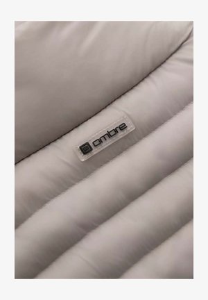 Hellgrauer Pufferstoff mit genähten Abschnitten, glatter Textur und einem Plastik-Tag mit dem Wort "ombre" in schwarzen Buchstaben.