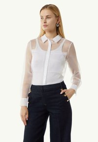 Weiße Bluse mit durchsichtigen langen Ärmeln und einer durchgehenden Knopfleiste, mit rundem Kragen und zwei Brusttaschen, kombiniert mit dunklen Jeanshosen.