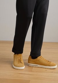 Gule lave sneakers med en teksturert overdel, hvite såler og runde lisser, kombinert med svarte oppbrettede bukser og en bakgrunn av tregulv.